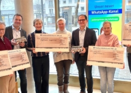 Es stehen 8 Personen an einem Schaufenster und halten große Checks einer Bank hoch.