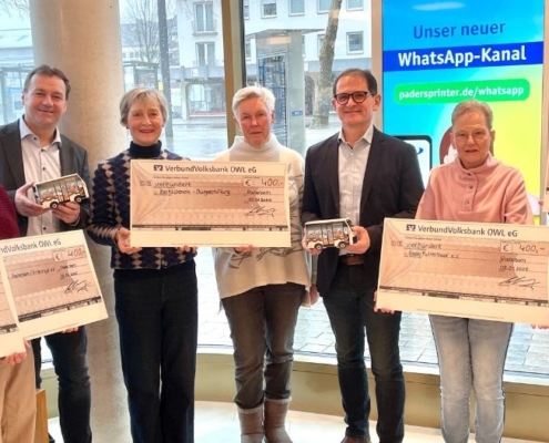 Es stehen 8 Personen an einem Schaufenster und halten große Checks einer Bank hoch.