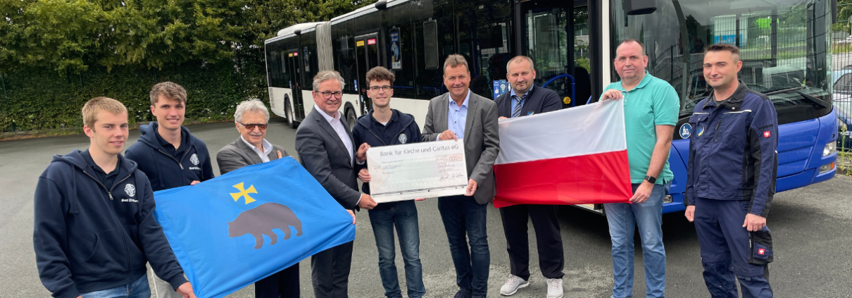 Neun Personen stehen vor einem Bus und präsentieren zwei Flaggen sowie einen Spendencheck