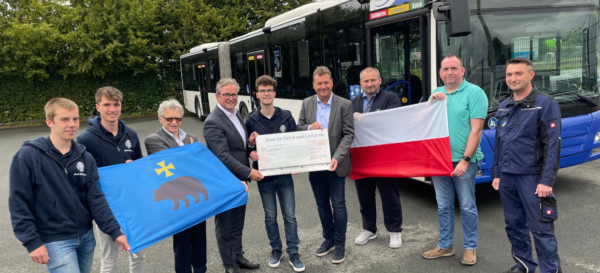 Neun Personen stehen vor einem Bus und präsentieren zwei Flaggen sowie einen Spendencheck