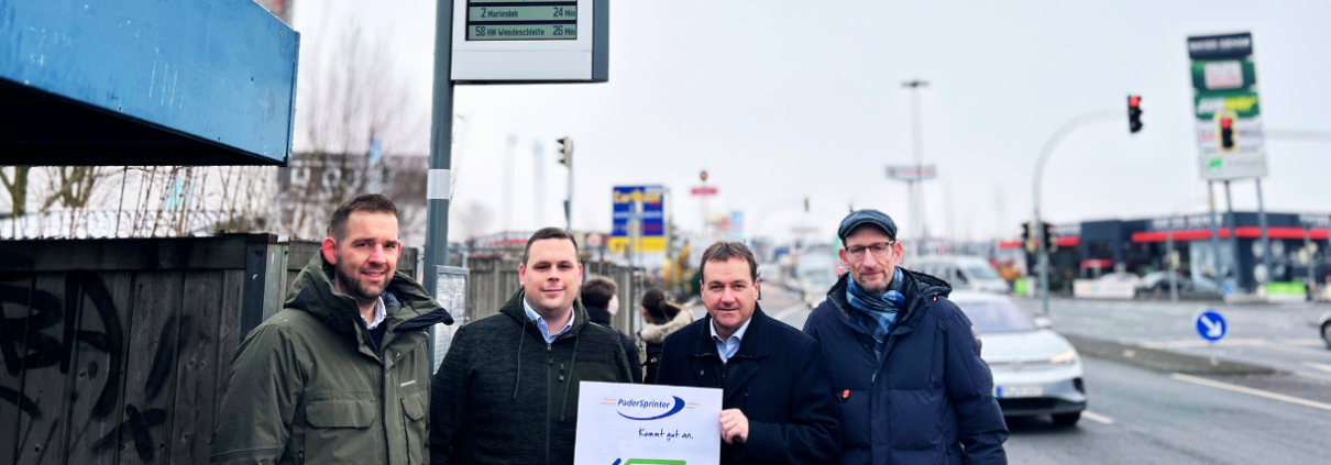 Zwei Mitarbeiter des PaderSprinters sowie zwei Mitarbeiter des NWL stehen vor einem Haltestellenschild mit einer dynamischen Fahrgastinformationsanzeige