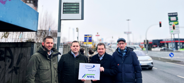 Zwei Mitarbeiter des PaderSprinters sowie zwei Mitarbeiter des NWL stehen vor einem Haltestellenschild mit einer dynamischen Fahrgastinformationsanzeige