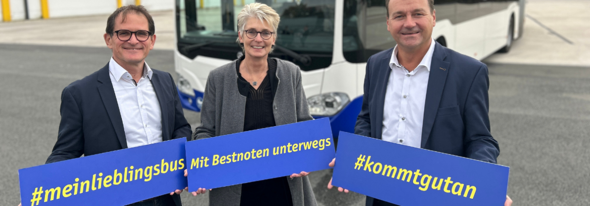 Drei Personen stehen vor einem Gelenkbus und zeigen jeweils ein Schild mit den Aufschriften "#meinlieblingsbus", "Mit Bestnoten unterwegs" sowie "#kommtgutan"