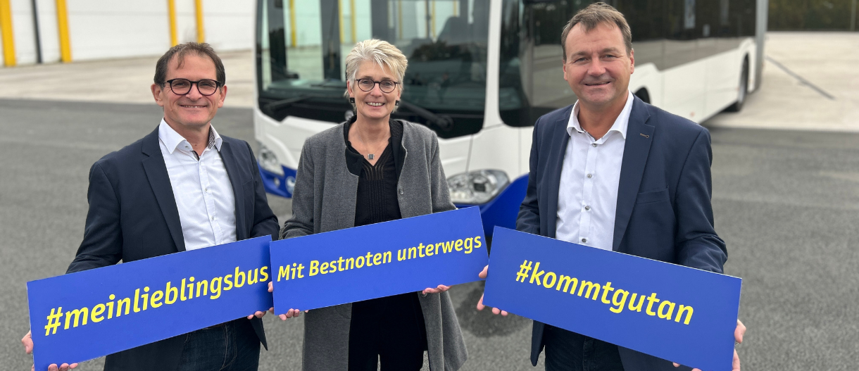 Drei Personen stehen vor einem Gelenkbus und zeigen jeweils ein Schild mit den Aufschriften "#meinlieblingsbus", "Mit Bestnoten unterwegs" sowie "#kommtgutan"