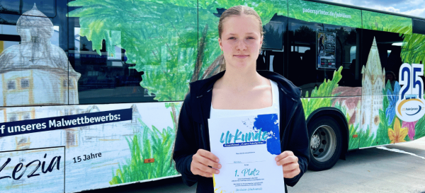 Ein junges Mädchen steht vor einem Bus mit einem bunten, selbst gemalten Design und hält stolz ihre Siegerurkunde in den Händen. Im Hintergrund ist das Kunstwerk im Dschungel-Look auf dem Bus zu sehen.