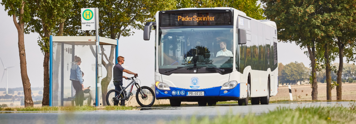 Ein Bus des PaderSprinter hält an einer Bushaltestelle, um dort mehrere Personen einsteigen zu lassen, davon eine Person mit einem Fahrrad