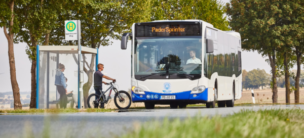 Ein Bus des PaderSprinter hält an einer Bushaltestelle, um dort mehrere Personen einsteigen zu lassen, davon eine Person mit einem Fahrrad
