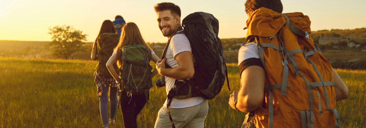 Fünf Personen wandern bei Sonnenschein durch eine Wiese, während eine Person lächelnd ins Bild schaut