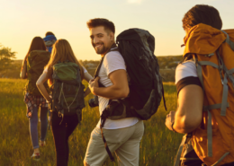 Fünf Personen wandern bei Sonnenschein durch eine Wiese, während eine Person lächelnd ins Bild schaut