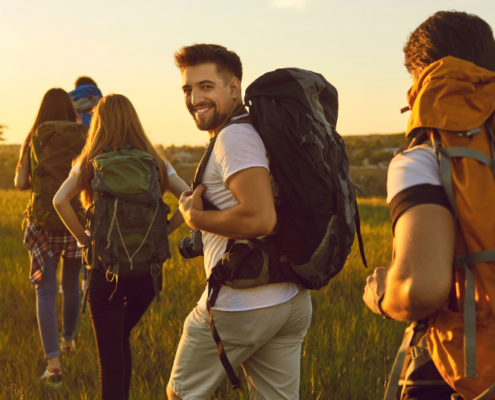 Fünf Personen wandern bei Sonnenschein durch eine Wiese, während eine Person lächelnd ins Bild schaut