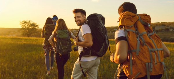 Fünf Personen wandern bei Sonnenschein durch eine Wiese, während eine Person lächelnd ins Bild schaut