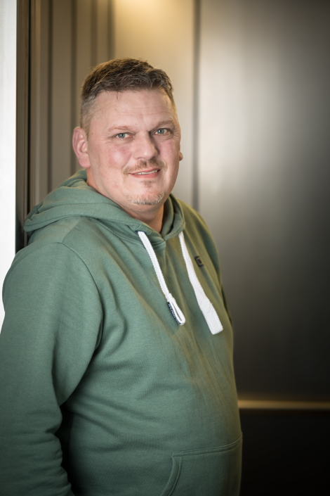 Portraitfoto von Carsten, Facility Manager bei PaderSprinter: lächelnd in grünem Kapuzenpullover, lehnt entspannt an den Fahrstuhl im Betriebsgebäude.