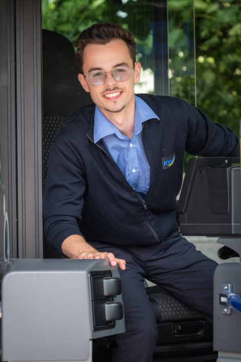 Portraitfoto von Fabian, Auszubildender zur Fachkraft im Fahrbetrieb bei PaderSprinter: junger Mann mit Brille und Dienstkleidung sitzt lächelnd im Busfahrerarbeitsplatz und blickt aus der offenen Tür.
