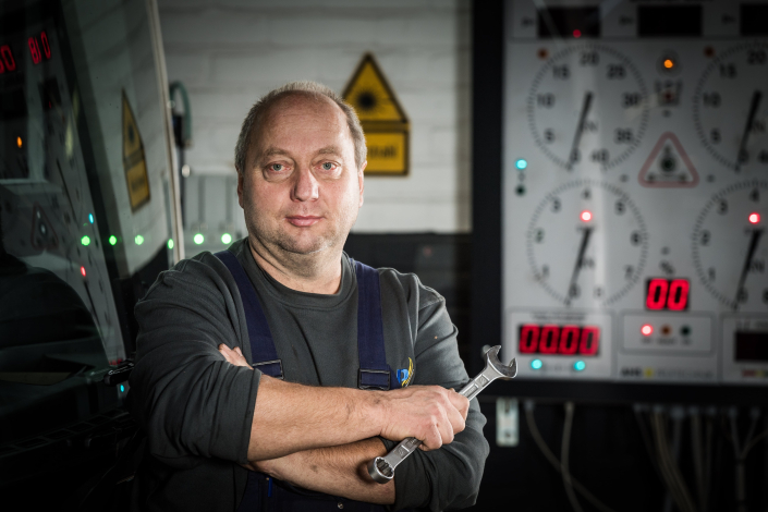 Portraitfoto von Heinrich, Kfz‑Mechaniker bei PaderSprinter: steht mit verschränkten Armen und Ringmaulschlüssel vor einem Prüfstand in der Werkstatt, daneben spiegelt sich ein Bus.