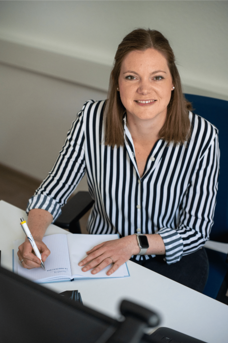 Portraitfoto von Jennifer, Marketingleiterin bei PaderSprinter: sitzt lächelnd am Schreibtisch, trägt schwarz‑weiß gestreifte Bluse und schreibt Notizen in ein offenes Heft.
