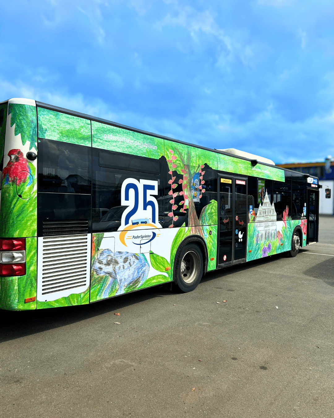 Gesamte Seitenansicht des grün und bunt bemalten PaderSprinter Jubiläumsbusses, der auf einem Parkplatz steht, mit Bäumen, Vögeln, Blumen und der großen Zahl 25.