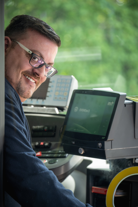 Portraitfoto von Mauro, Bus‑ und Lehrfahrer bei PaderSprinter: lächelt über die Schulter aus dem Fahrerplatz, trägt Brille und sitzt vor Armaturenbrett mit Ticketterminal.