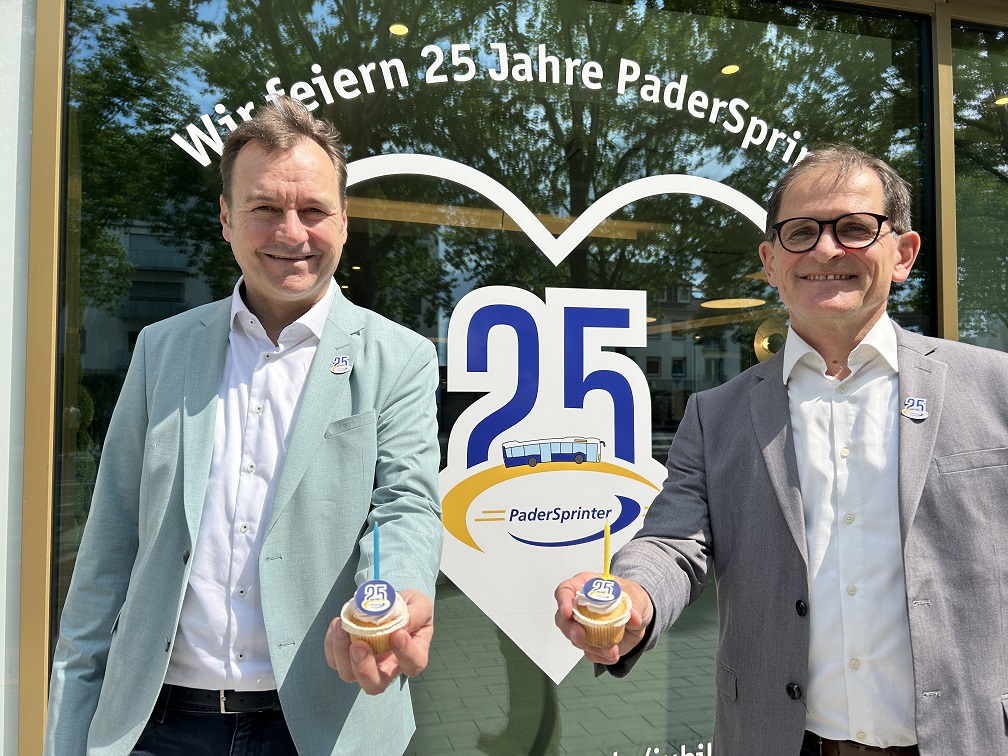 Der Geschäftsführer des PaderSprinter, Oliver Eikenberg, und der kaufmännische Leiter, Andreas Frank, stehen vor dem Kundencenter. Sie haben einen Cupcake in der Hand. Im Hintergrund erkennt man an der Scheibe "Wir feiern 25 Jahre PaderSprinter"