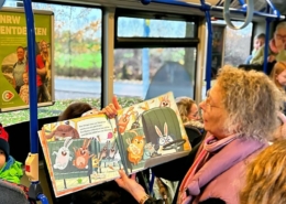 Meherer Kinder sitzen im Bus. Eine Erwachsene Frau hält ein Buch hoch und sagt etwas dazu. Alle hören ihr zu.