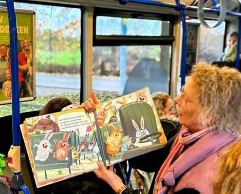 Meherer Kinder sitzen im Bus. Eine Erwachsene Frau hält ein Buch hoch und sagt etwas dazu. Alle hören ihr zu.